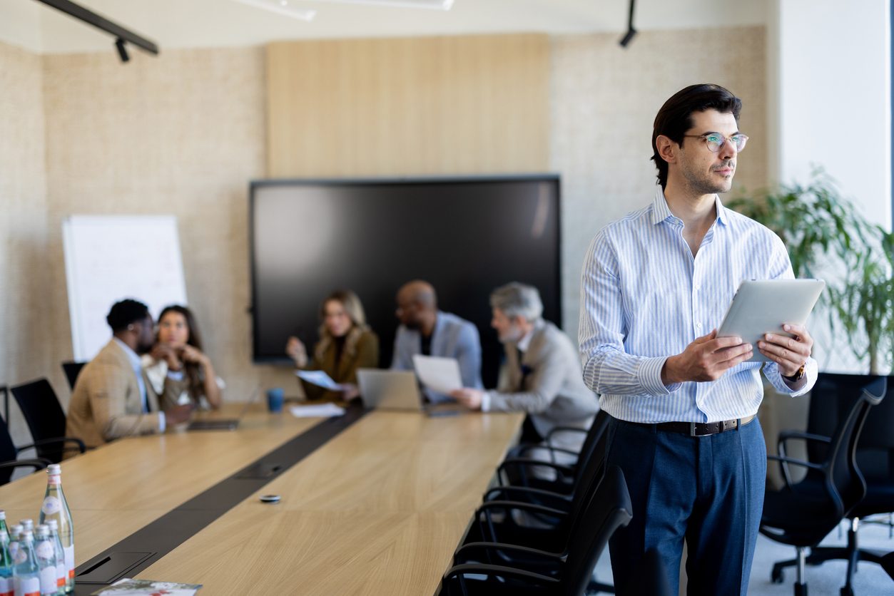 An executive holding a tablet in a boardroom while colleagues meet at the conference table behind him