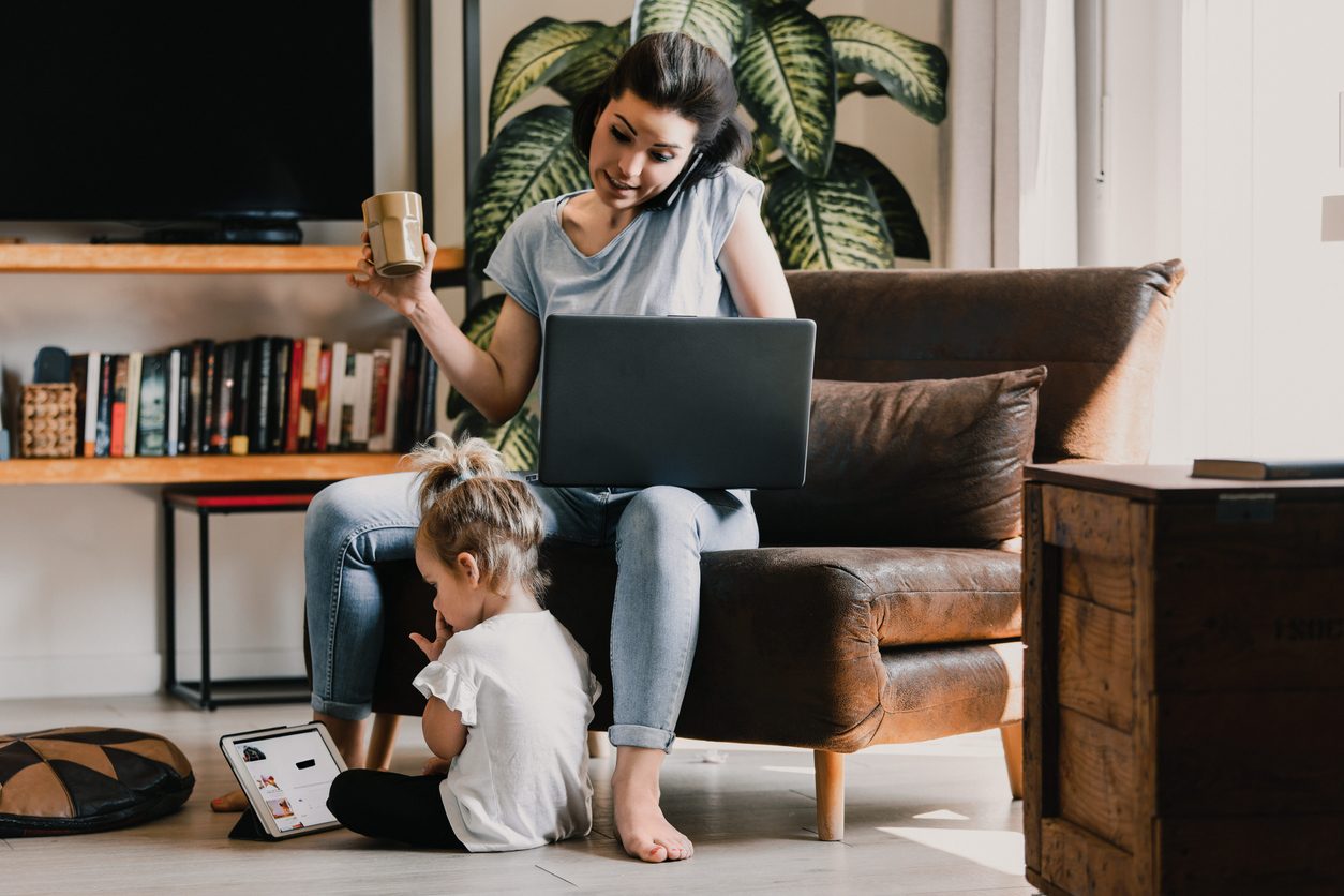 A mother working from home on a laptop and phone while her young child plays on the floor beside her